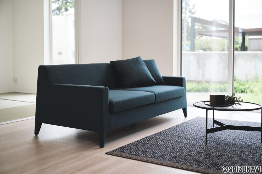 【B棟】クリーンなラインと無駄のないデザインが特徴のソファーは深みのあるブルー系をセレクト。ソファーと合わせて、ラグとアクセントクロスもブルー系をコーディネートしてリビング全体に統一感をだしました。