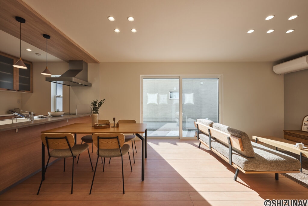 【B棟】大きな窓から光がたっぷり差し込む、のびやかなLDK。木目のキッチンと家具がやさしく調和し、食事もくつろぎも家族の時間が自然と深まる空間です。